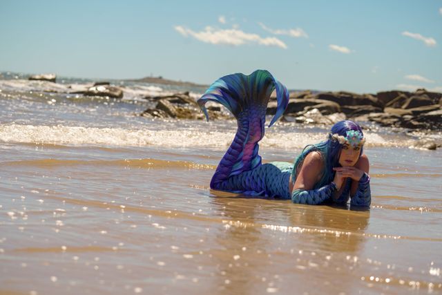Kaz lying in the sunlit shallows of a Tasmanian beach in her blue and violet mermaid tail
