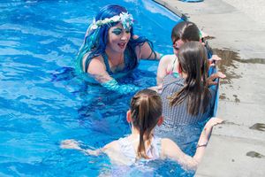 Kaz in the pool as a mermaid, chatting with children at the pool edge