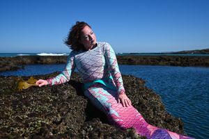 Kaz reclining on coastal rocks beside a rockpool, wearing a pink and aqua mermaid tail