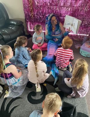 Kaz in her blue mermaid tail reading a story to a circle of children at a birthday party